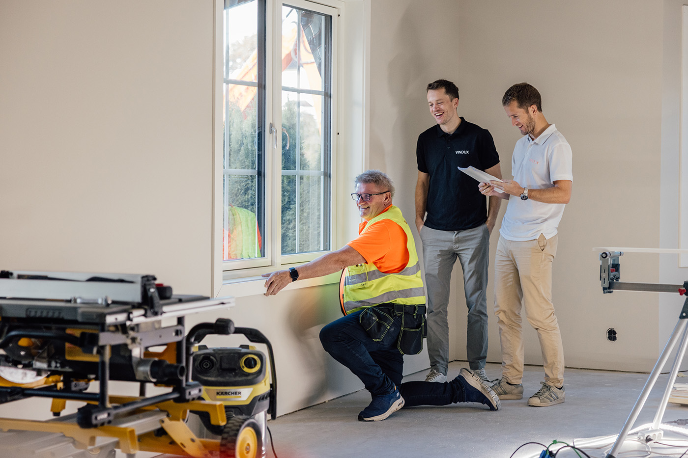 En bygningsarbeider monterer lister ved et vindu, mens to menn står i nærheten, den ene med en skriveplate i hånden. Verktøy og utstyr er synlig i rommet.