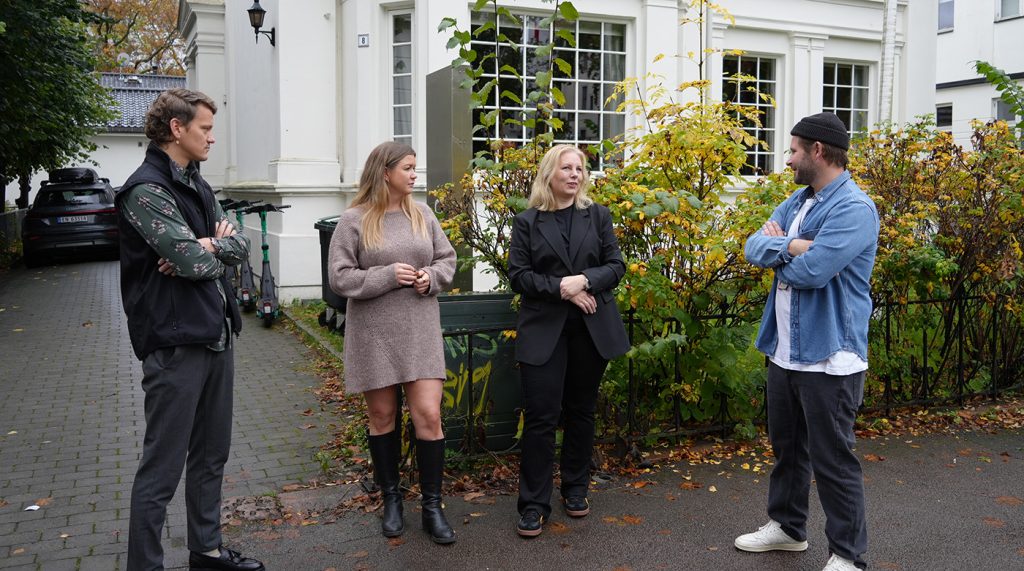 Fire personer står utenfor en hvit bygning og snakker sammen. To kvinner står i midten, flankert av to menn. Scenen utspiller seg på en asfaltert sti med høstløv.