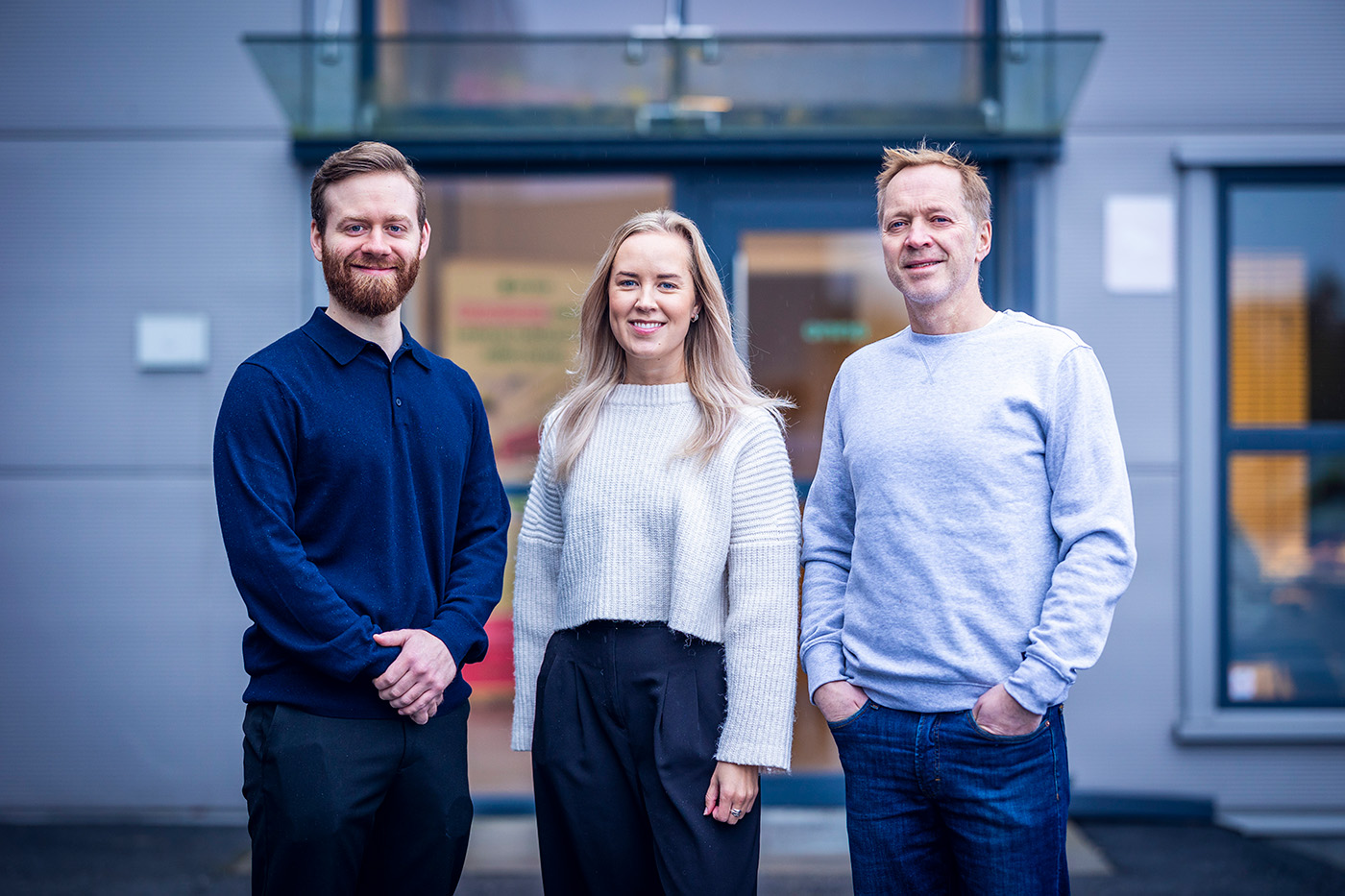 Tre personer står side om side utenfor en moderne bygning, vendt mot kameraet og smiler.