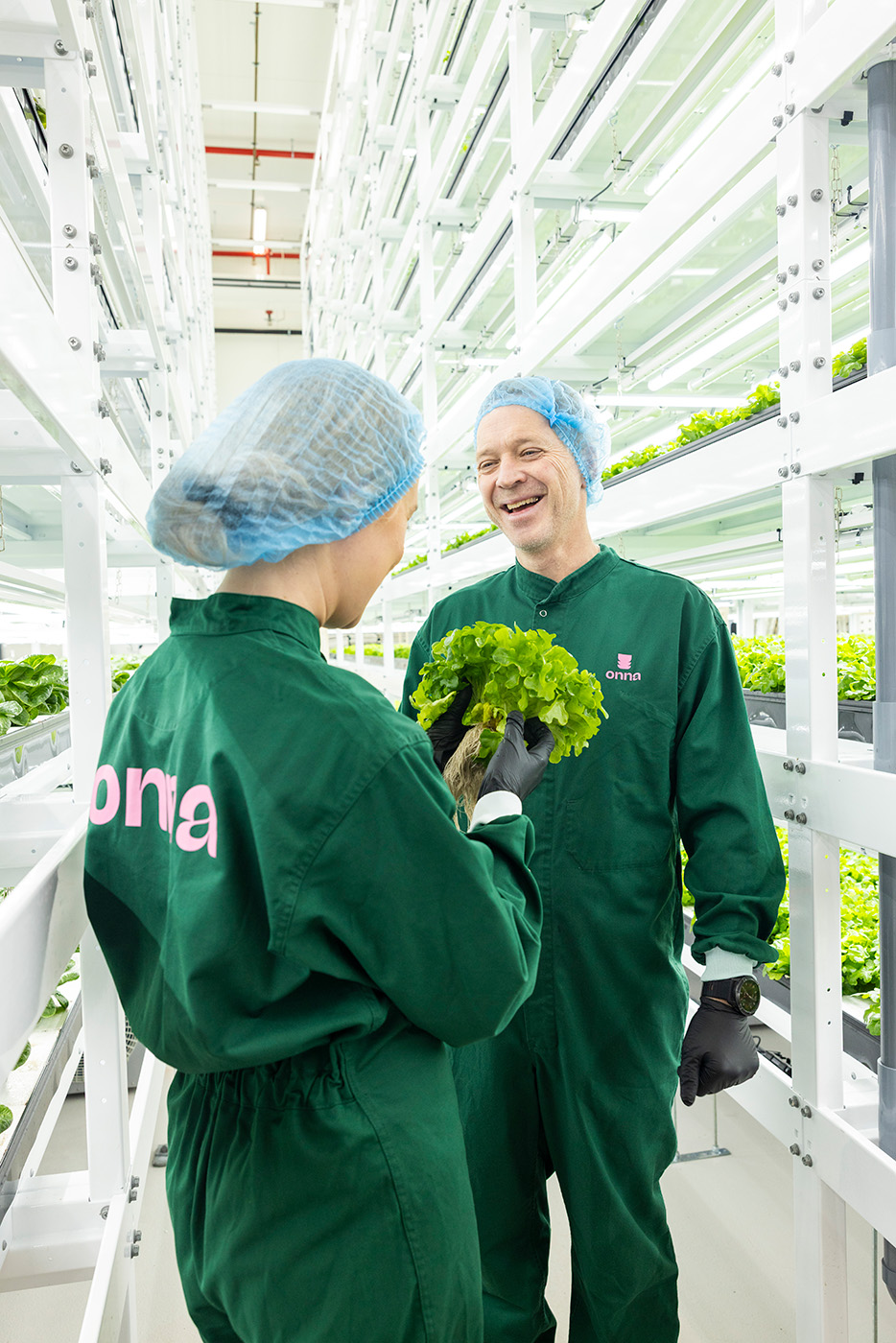 To personer i grønne uniformer og hårnett står i en vertikal innendørs gård og holder og diskuterer et hode med bladgrønn salat.