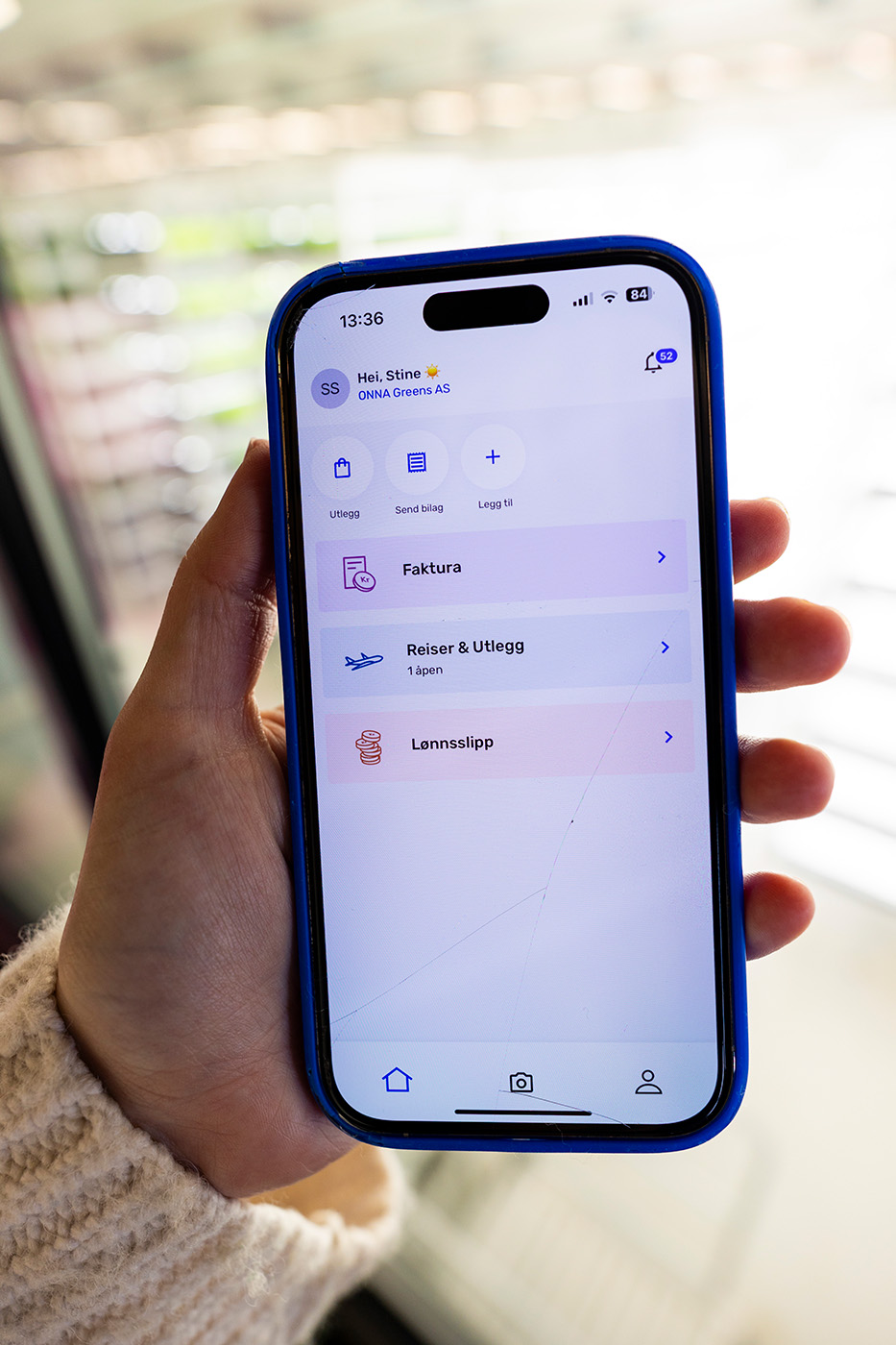 En hånd som holder en smarttelefon som viser en økonomi- eller forretningsapp med alternativer for fakturaer, reiseregninger og lønnsslipper.