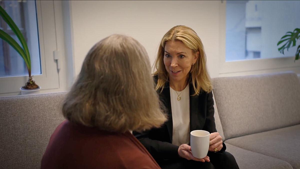 To kvinner sitter i en sofa på et kontor og snakker sammen, den ene holder et hvitt krus.