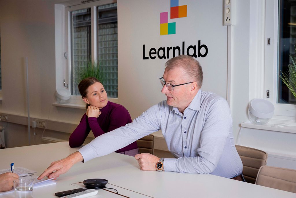 To personer sitter ved et konferansebord i et møterom med Learnlab-logoen på veggen; mannen peker på et dokument på bordet.