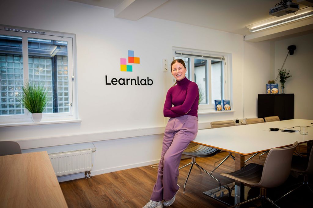 En kvinne i lilla antrekk står smilende ved et konferansebord i et moderne kontor med en "Learnlab"-logo på veggen.