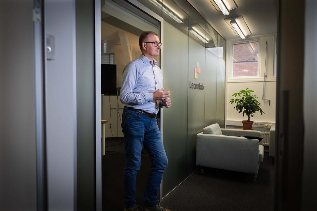 En mann står i et moderne kontor ved siden av en glassvegg med "Learnlab"-logoen, i nærheten av en sofa og en potteplante.