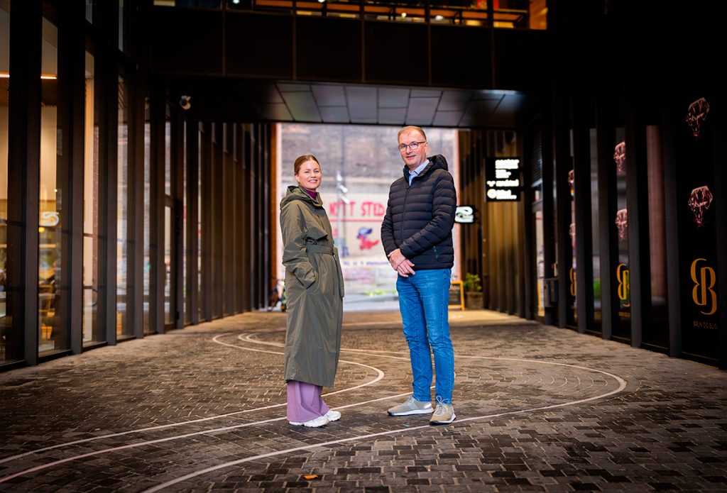 To personer står på en asfaltert gangvei mellom moderne bygninger, vendt mot kameraet. Kvinnen har på seg en grønn frakk, mannen en mørk jakke og jeans.