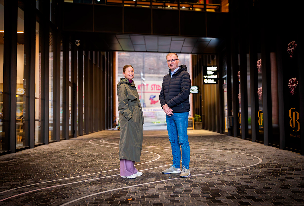 To personer står på en asfaltert gangvei mellom moderne bygninger, vendt mot kameraet. Kvinnen har på seg en grønn frakk, mannen en mørk jakke og jeans.