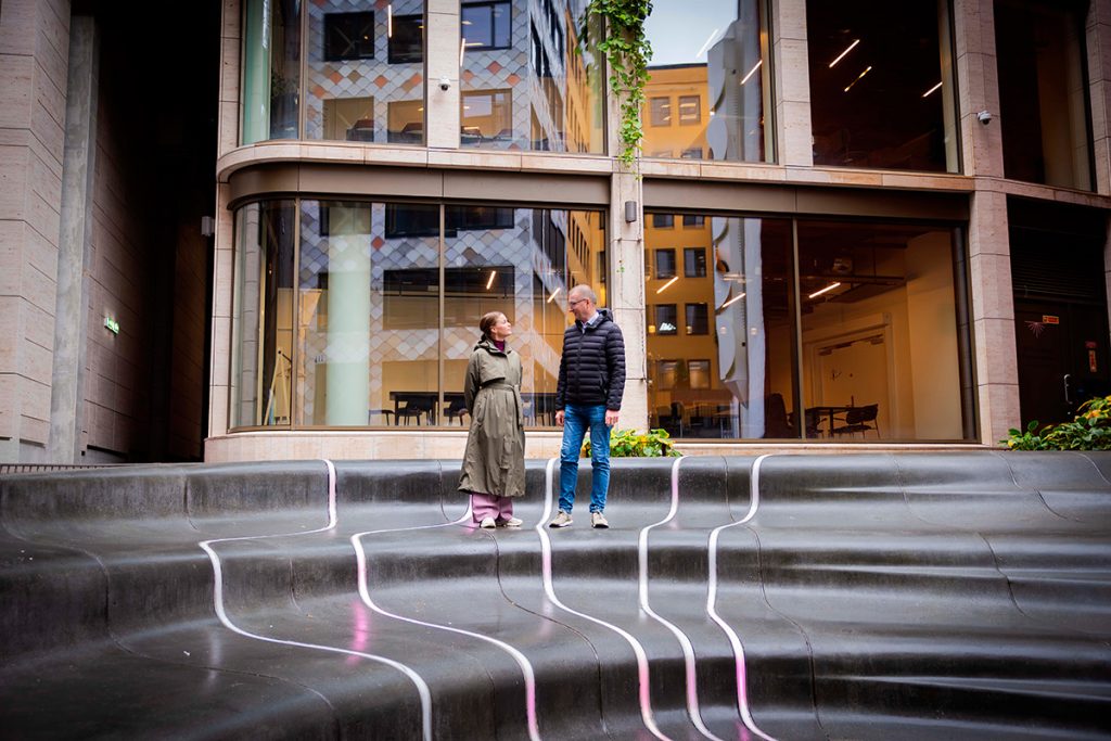 To personer står på sorte trappetrinn med hvite linjer foran en moderne glassbygning.