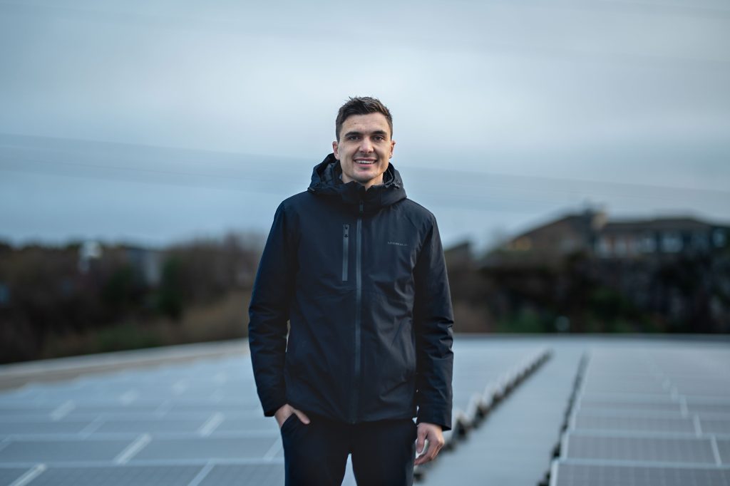 En mann i mørk jakke står på et hustak med solcellepaneler, med en uskarp bakgrunn av trær og bygninger på en overskyet dag.