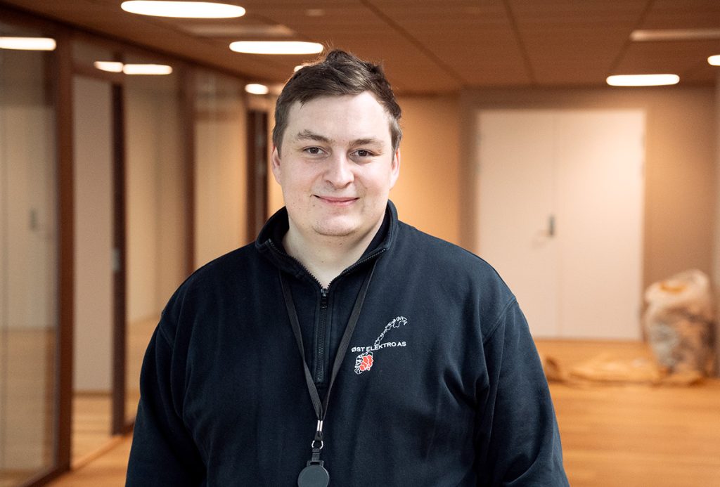 En person i svart genser og nøkkelbånd står i en moderne gang med kontorer med glassvegger og tregulv.