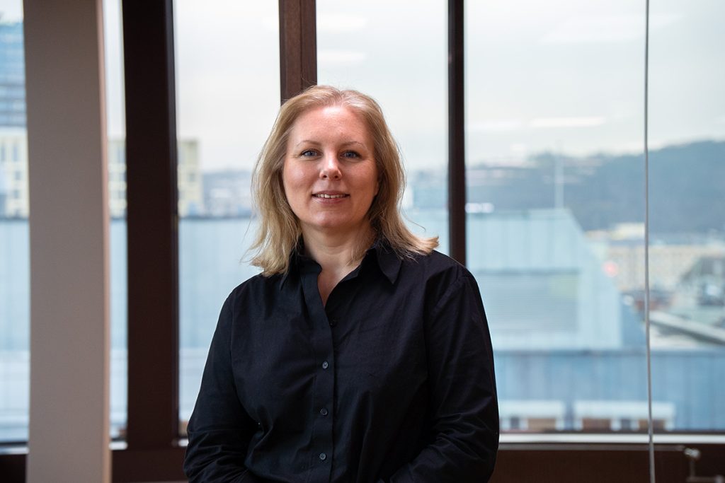 En kvinne med skulderlangt, blondt hår og svart skjorte står innendørs foran store vinduer med et bylandskap i bakgrunnen.