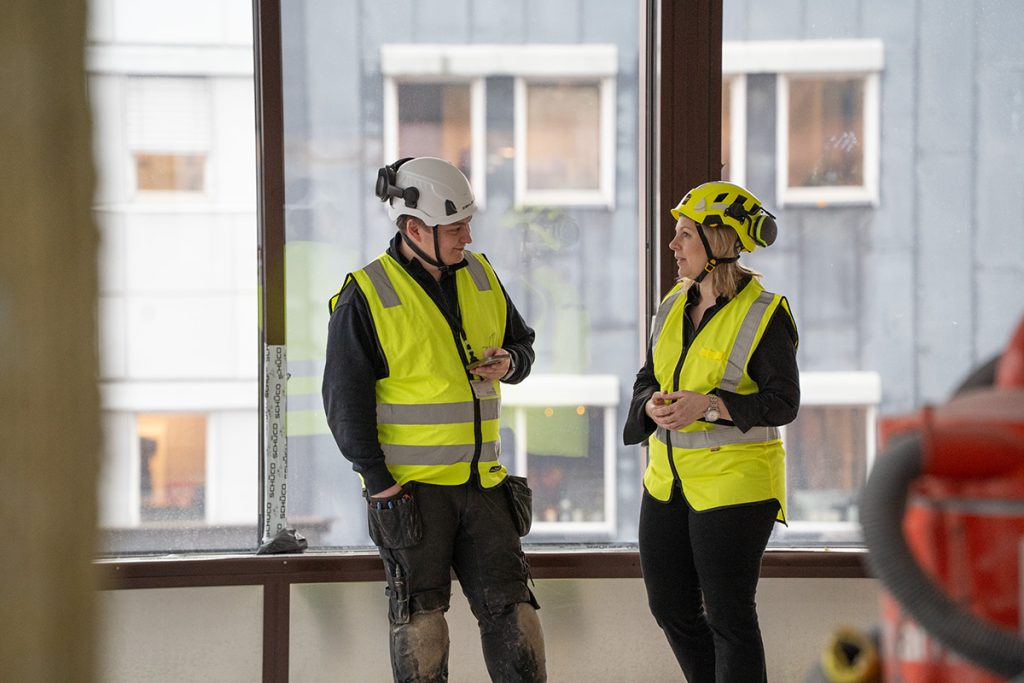 To personer iført vernehjelm og varselvest står innendørs på en byggeplass og snakker sammen i nærheten av store vinduer.