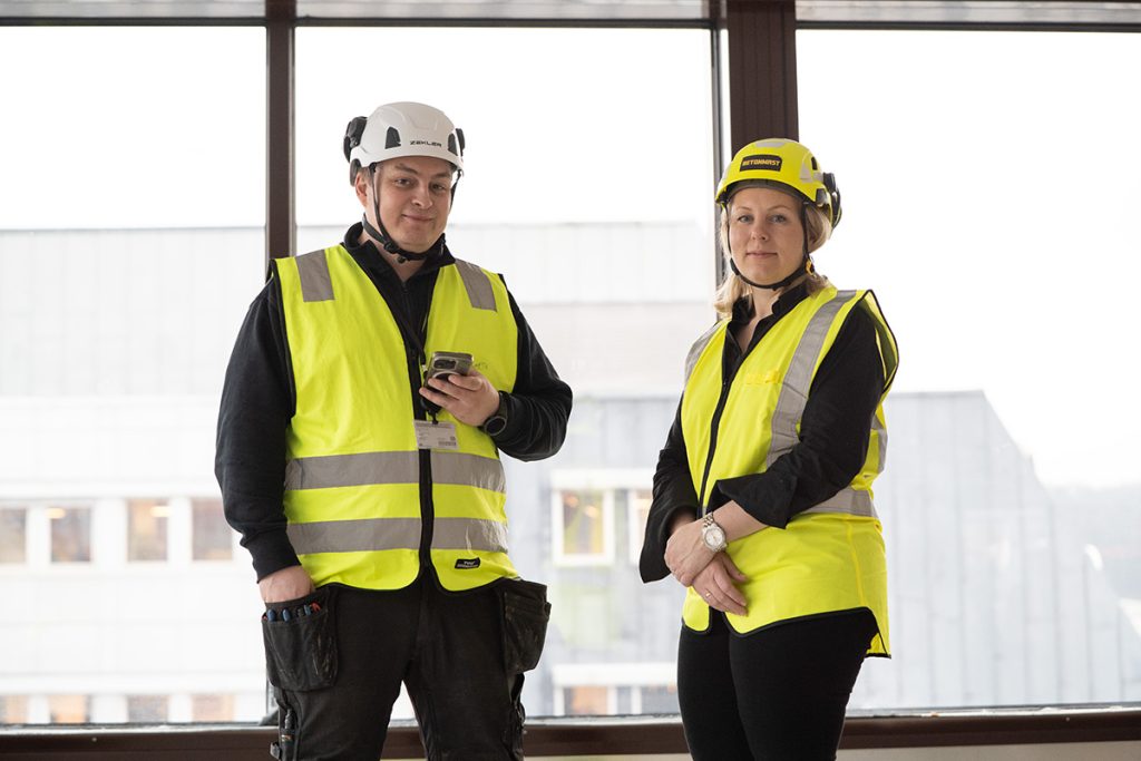 To bygningsarbeidere iført gule sikkerhetsvester og hjelmer står innendørs ved store vinduer, den ene med en telefon i hånden.