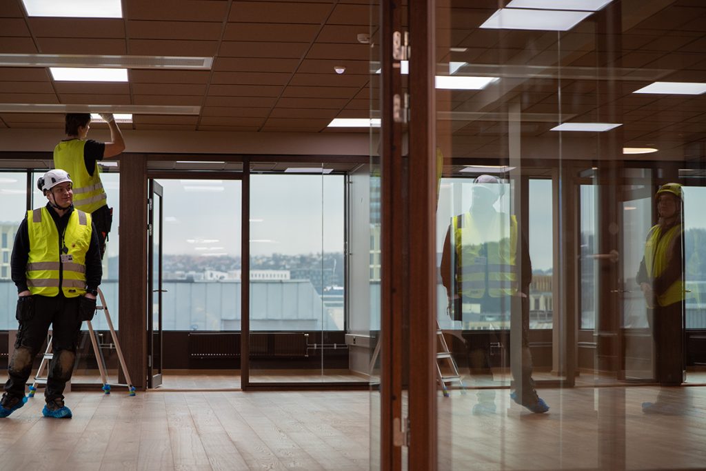 To arbeidere i gule vernetrøyer og hjelmer pusser vinduer i et moderne kontor med glassvegger og utsikt over byen.