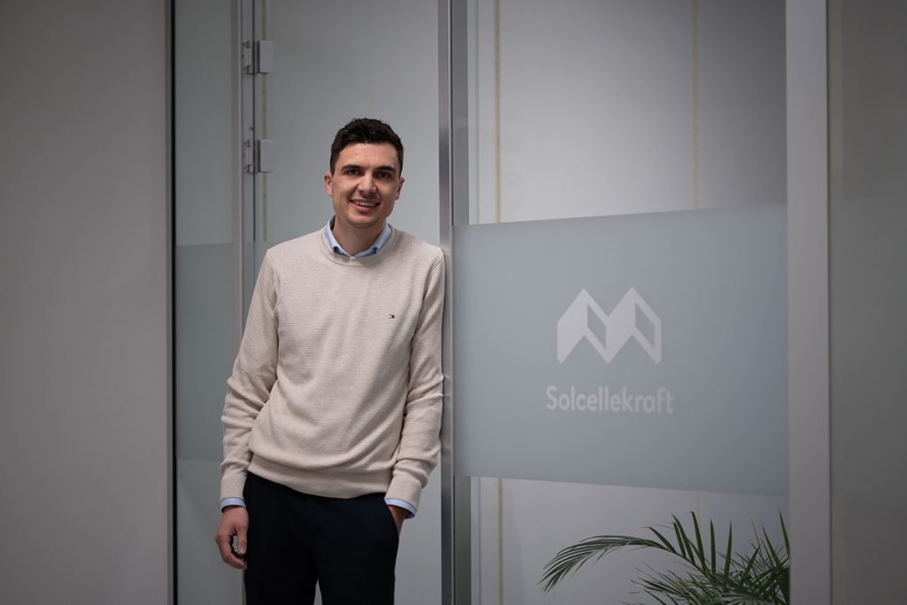 En mann i lys genser står og smiler ved en kontordør i glass med logoen og teksten "Solcellekraft".