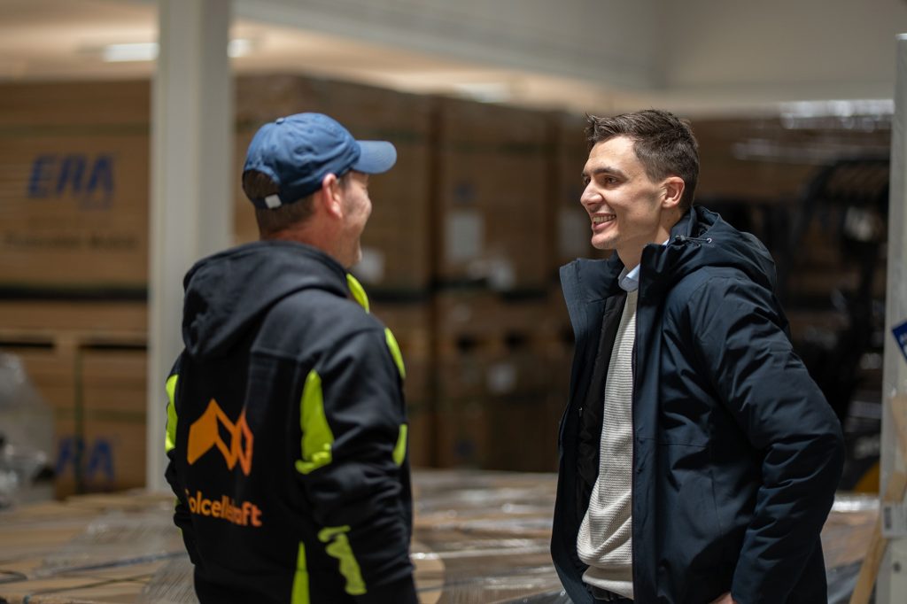 To menn står og smiler og snakker sammen i en lagerbygning, med stablede esker synlige i bakgrunnen.