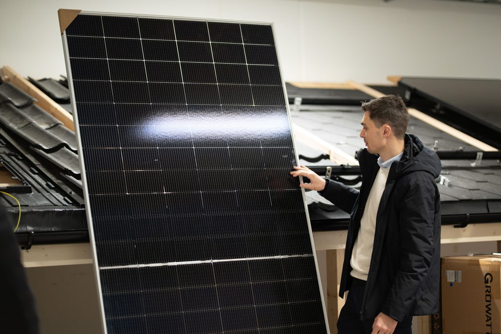 En person i svart jakke undersøker et stort solcellepanel innendørs, med ekstra utstyr og materialer synlig i bakgrunnen.