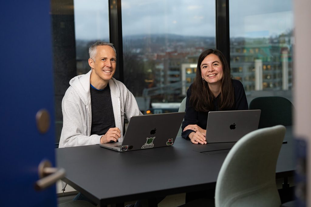 Vidar og Negina fra Tripletex' utviklingsavdeling sitter foran laptopene sine ved et bord på et moderne kontor og smiler til kameraet.