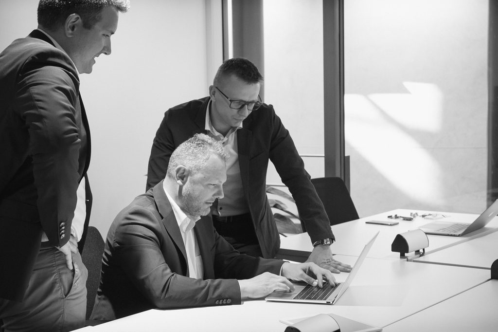 Tre menn i forretningsantrekk samles rundt et bord; én sitter og skriver på en bærbar datamaskin, mens de to andre står og ser på.
