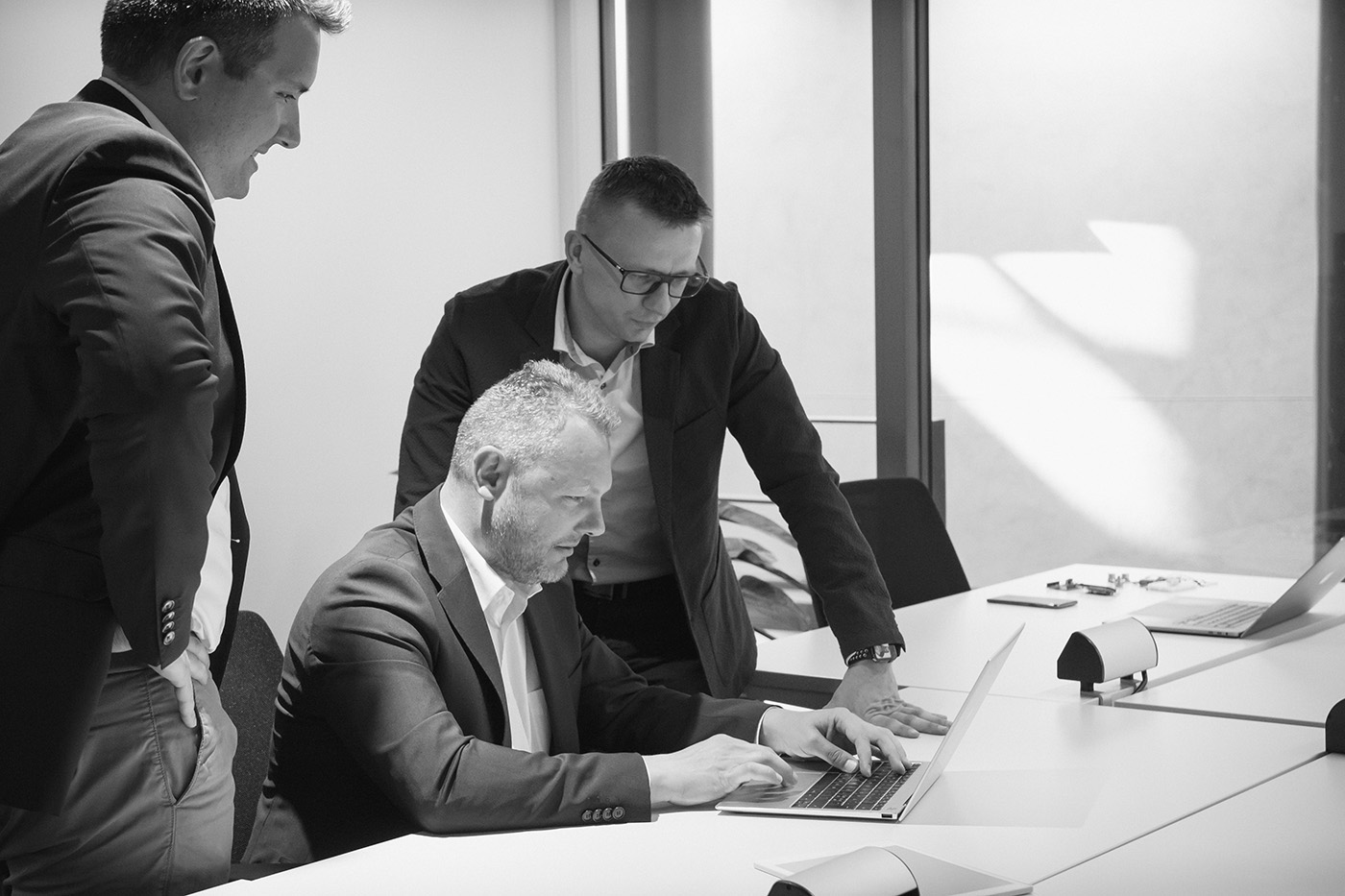 Tre menn i forretningsantrekk samles rundt et bord; én sitter og skriver på en bærbar datamaskin, mens de to andre står og ser på.