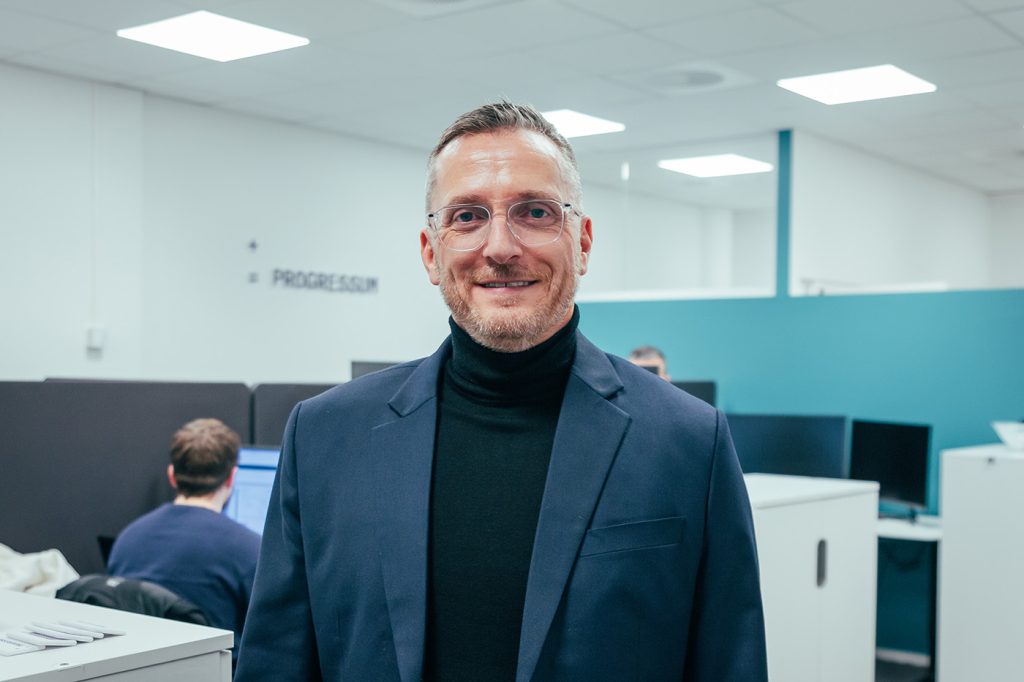 En mann med briller og marineblå blazer står på et kontor med pulter, datamaskiner og folk som jobber i bakgrunnen. Lars Gotaas i Progressum.