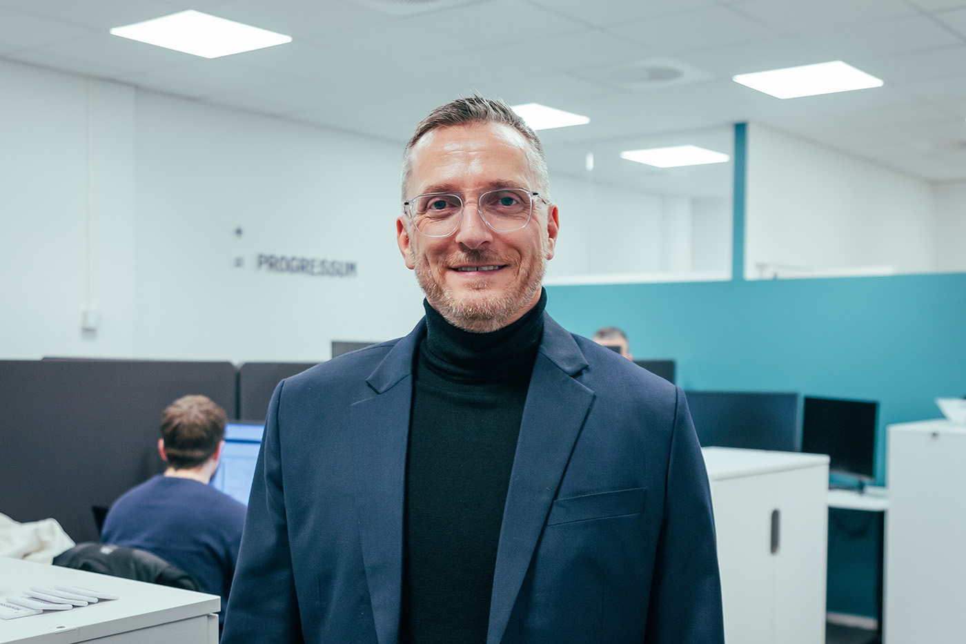 En mann med briller og marineblå blazer står på et kontor med pulter, datamaskiner og folk som jobber i bakgrunnen. Lars Gotaas i Progressum.