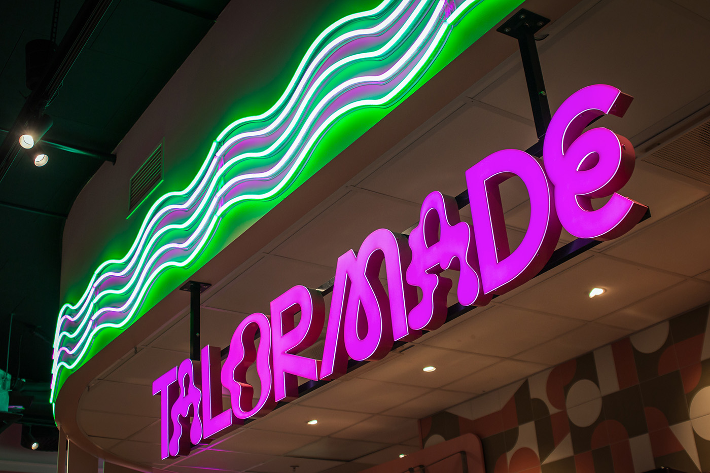 TAILORMADE" står det på et neonskilt med fete, rosa bokstaver og et bølgende, grønt neonlys over, inne i et moderne interiør.