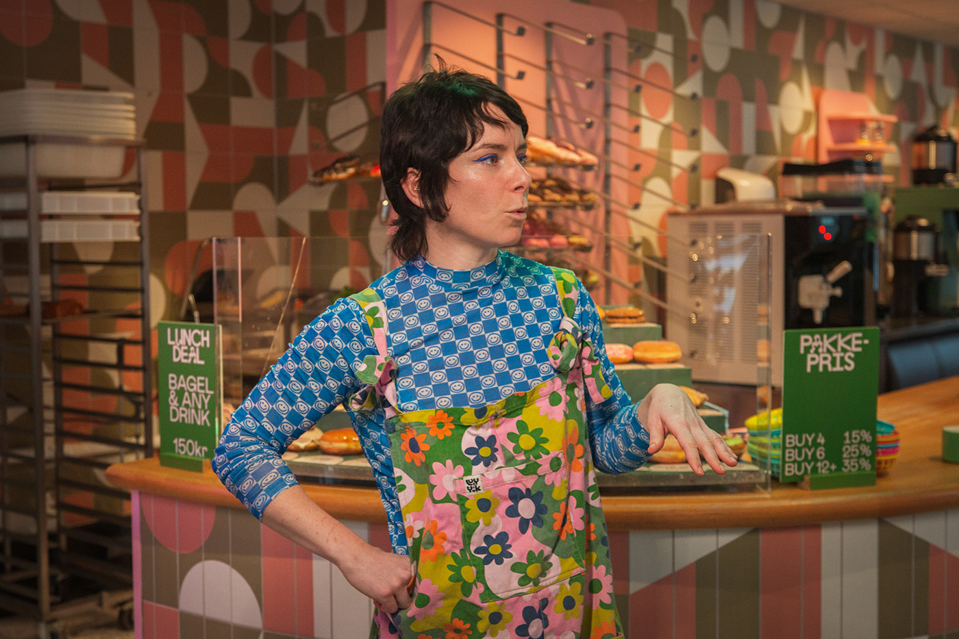 En person i et fargerikt, blomstrete forkle står i et bakeri og gestikulerer med den ene hånden. Bagels og reklameskilt er synlige i bakgrunnen på disken.