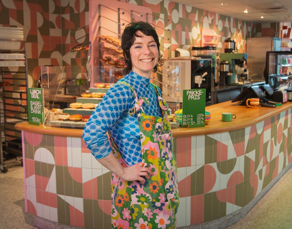 En kvinne i et fargerikt, blomstrete forkle står smilende foran en kafédisk med bakverk, drikke og menyskilt i bakgrunnen. Talor driver Talormade.