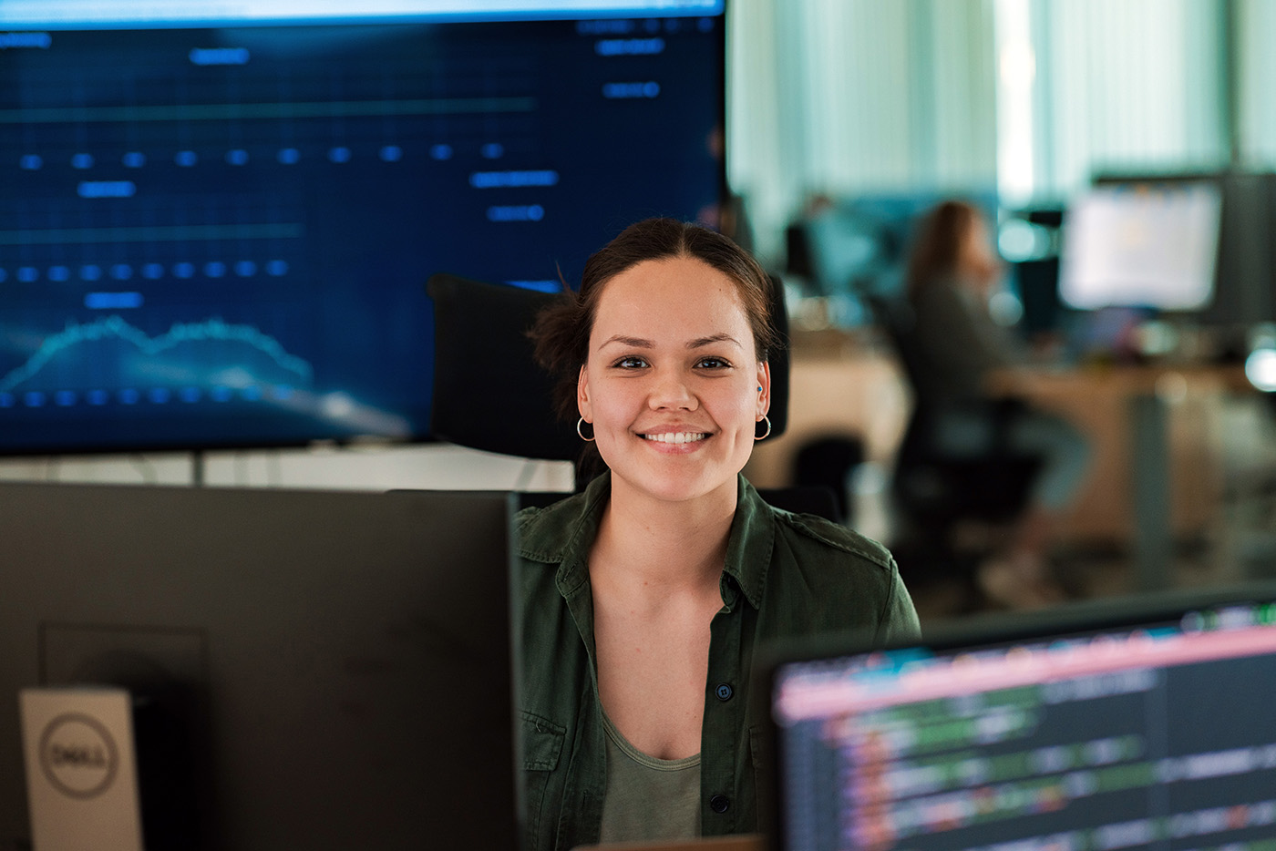 Kvinne smiler i kamera, sitter bak skjermer i et kontrolandskap