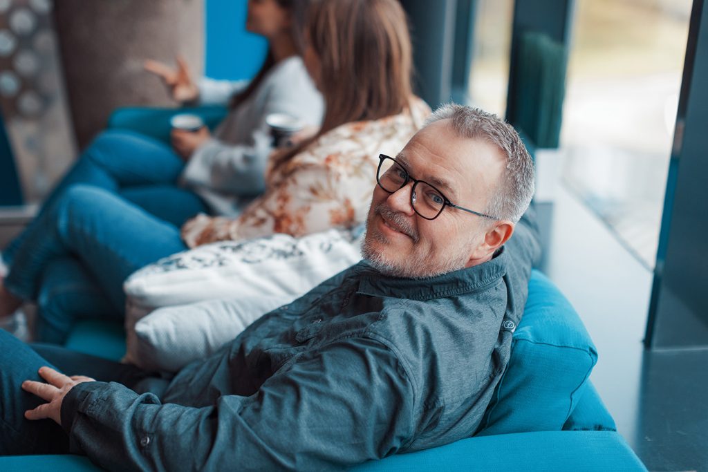 Mann med blå skjorte og briller sitter i en blå sofa, ser i kamera. To andre mennesker kan ses i bakgrunnen.