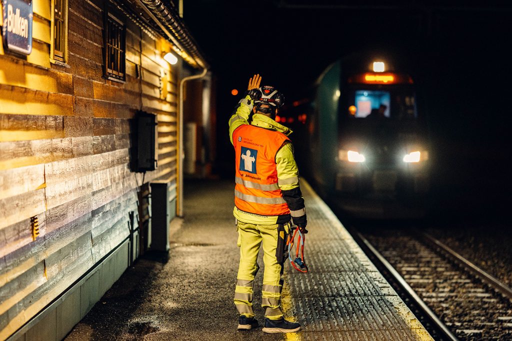 Anleggsarbeider står på perongen med ryggen til, vinker til et tog som ankommer perongen. Det er mørkt ute.