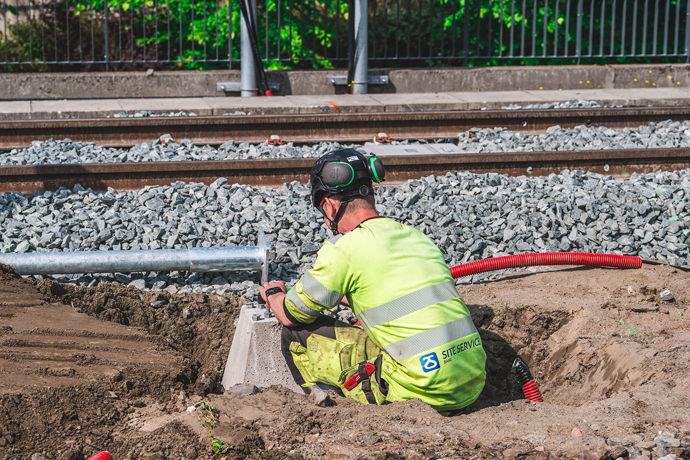 Mann fra Site Service Bane i gul arbeidsdress jobber på bakken, røde rør og togspor i bakgrunnen. 