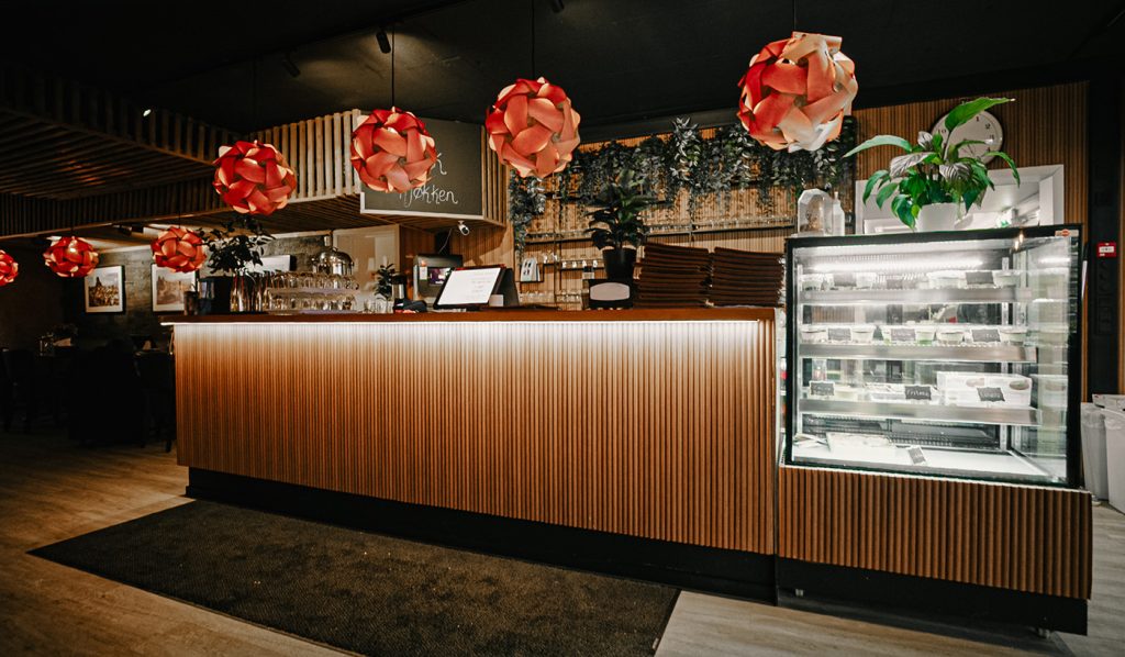 En moderne restaurantdisk med hengende røde pendellamper, en glassmonter fylt med desserter, potteplanter og stilig trepanel skaper en varm og innbydende restaurantatmosfære.
