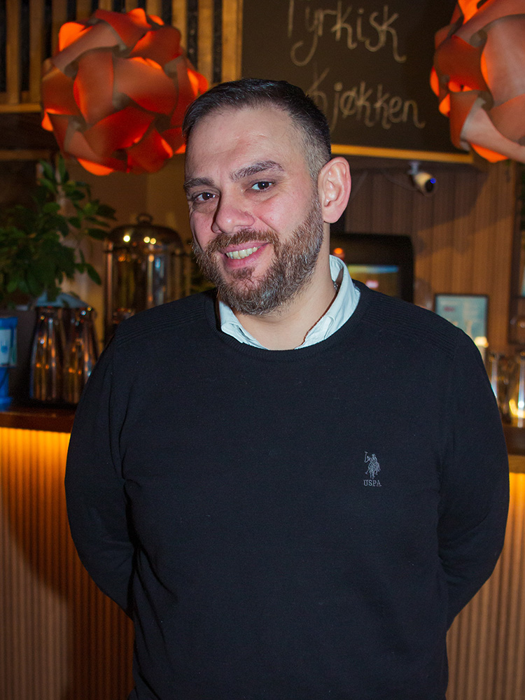Portrettbilde, mann med skjegg ser i kamera, står foran en bardisk i en restaurant.
