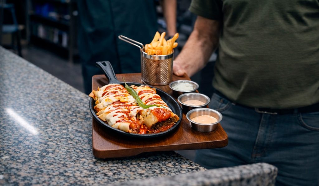 På en restaurant står en person med et trebrett med enchiladas toppet med saus og ost, en porsjon pommes frites i en metallkurv og tre dippesauser på en granittdisk.