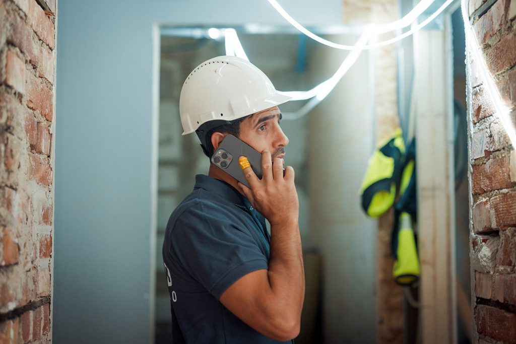 Mann prater i telefonen innendørs, på en byggeplass, lysslynge i bakgrunnen. Skal viderefakturere materiell for vvs- og elektro-jobb.