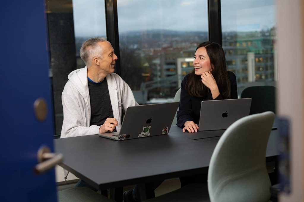 To personer fra Tripletex sitter ved et bord med laptoper, smiler og snakker sammen, med store vinduer og et bylandskap i bakgrunnen.