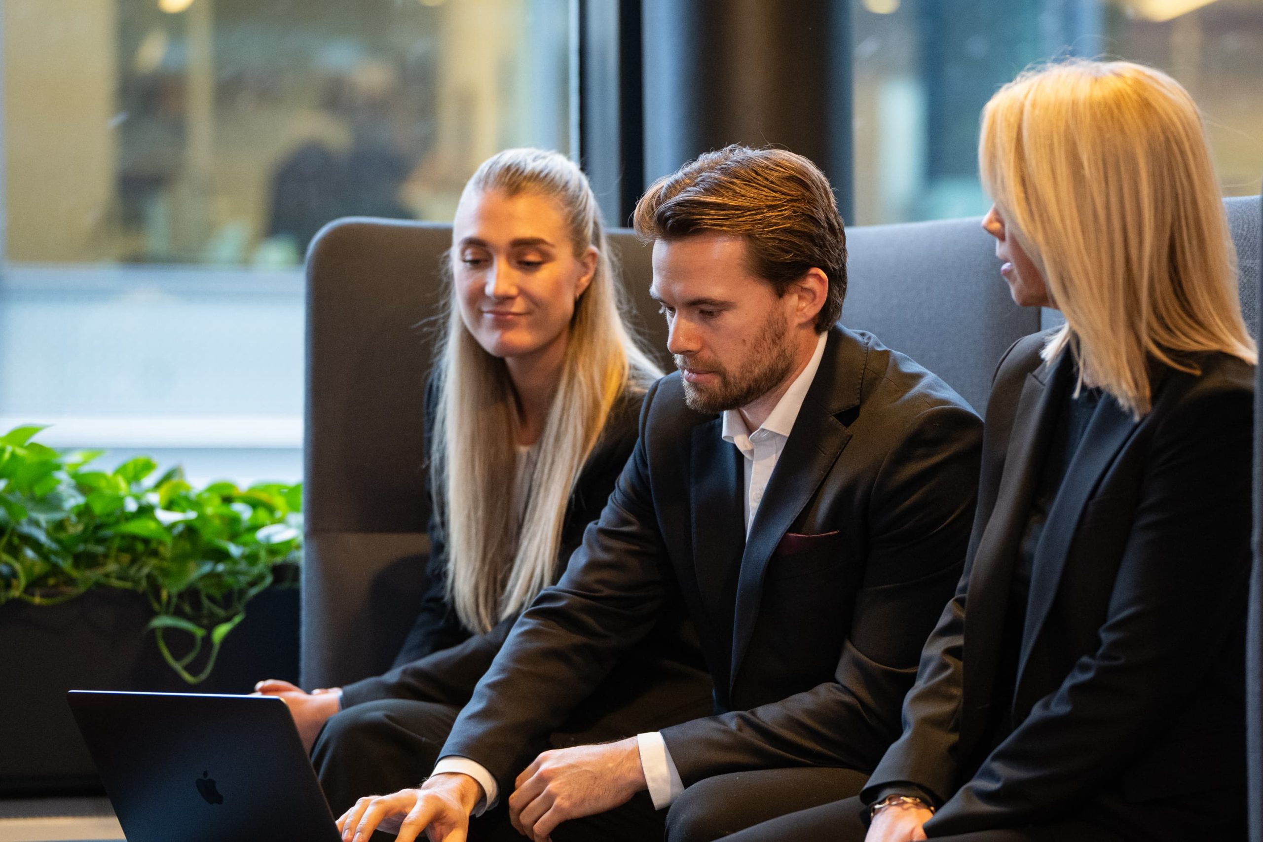 Tre personer i forretningsantrekk sitter på en benk og ser på en bærbar PC-skjerm sammen i et moderne kontor med vindu og planter, og diskuterer fordelene med et regnskapsprogram for advokater.