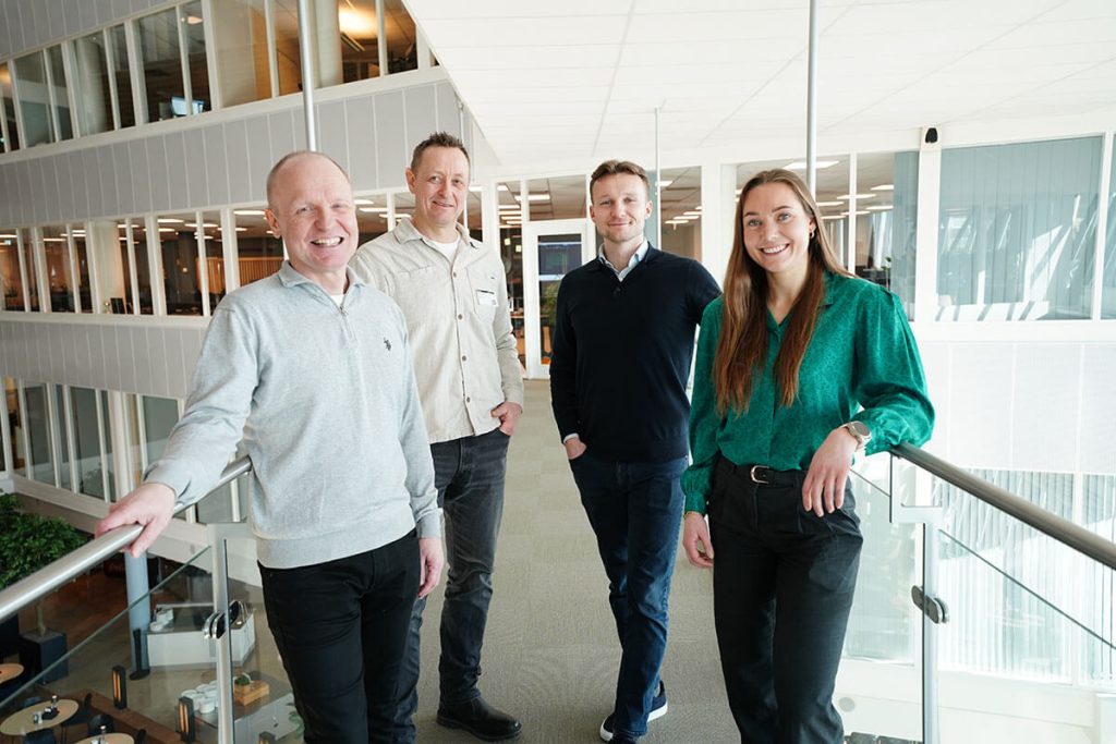 Fire personer, to fra selskapet Order Control og to fra Tripletex, står side om side i en moderne kontorgang og smiler til kameraet. Store vinduer og glassrekkverk er synlige i bakgrunnen.