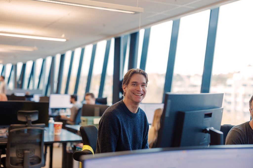 En mann sitter og smiler ved skrivebordet i et moderne kontorlokale med store vinduer og flere dataskjermer, selvsikker etter å ha tatt seg tid til å velge riktig regnskapsprogram for virksomheten sin.