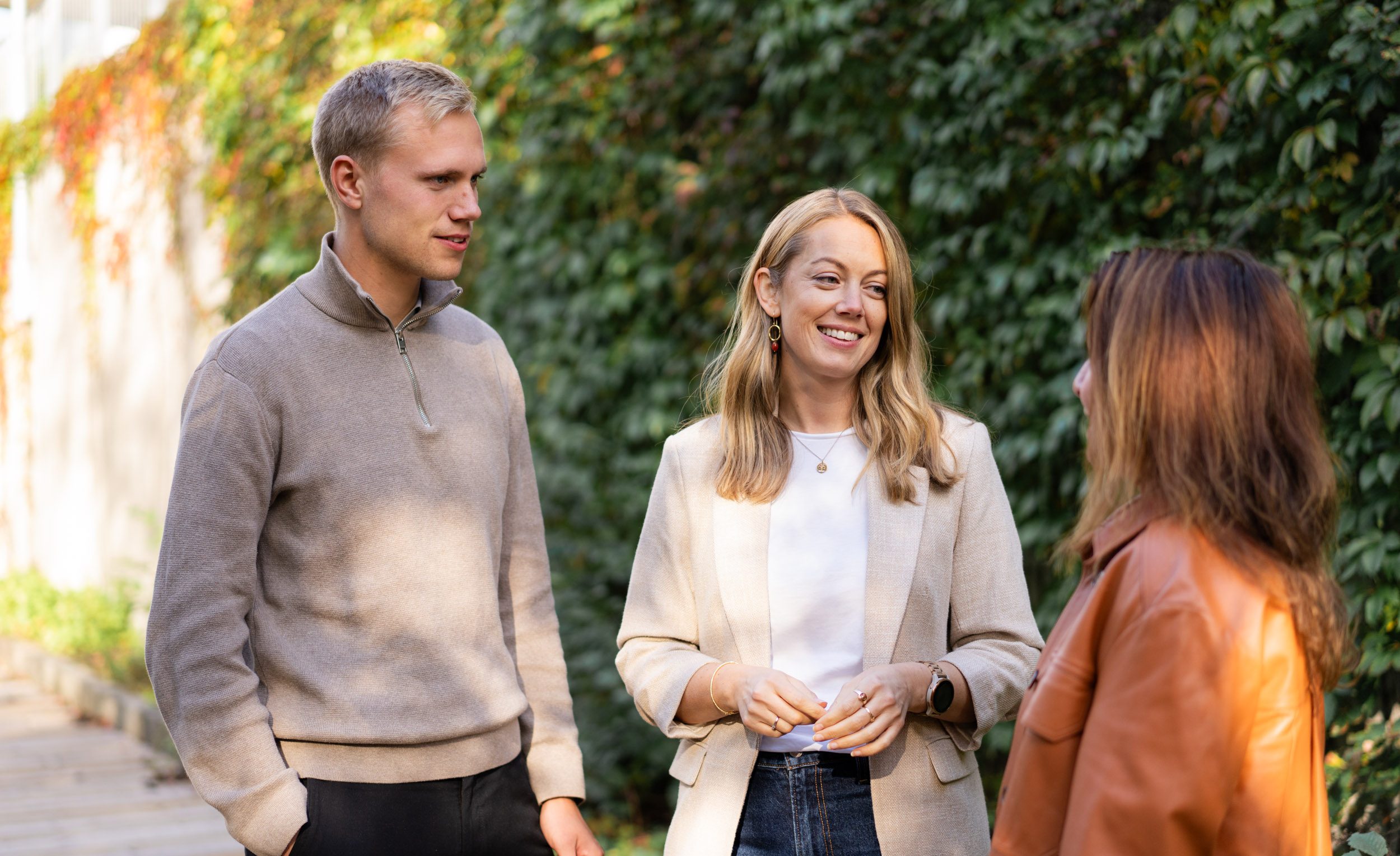 Tre personer står utendørs og snakker sammen. To kvinner og en mann er kledd i uformelle høstklær, og veggen bak dem er dekket av grønne planter.