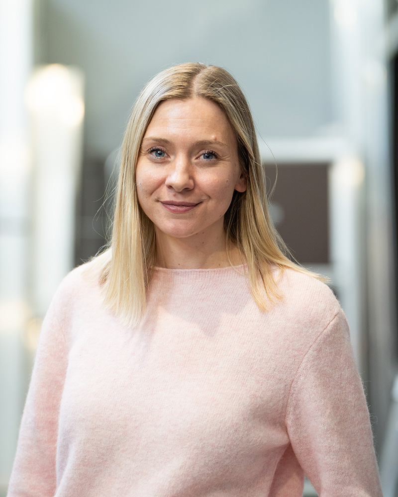 En kvinne med rett blondt hår, iført en lys rosa genser, står innendørs og smiler til kameraet.