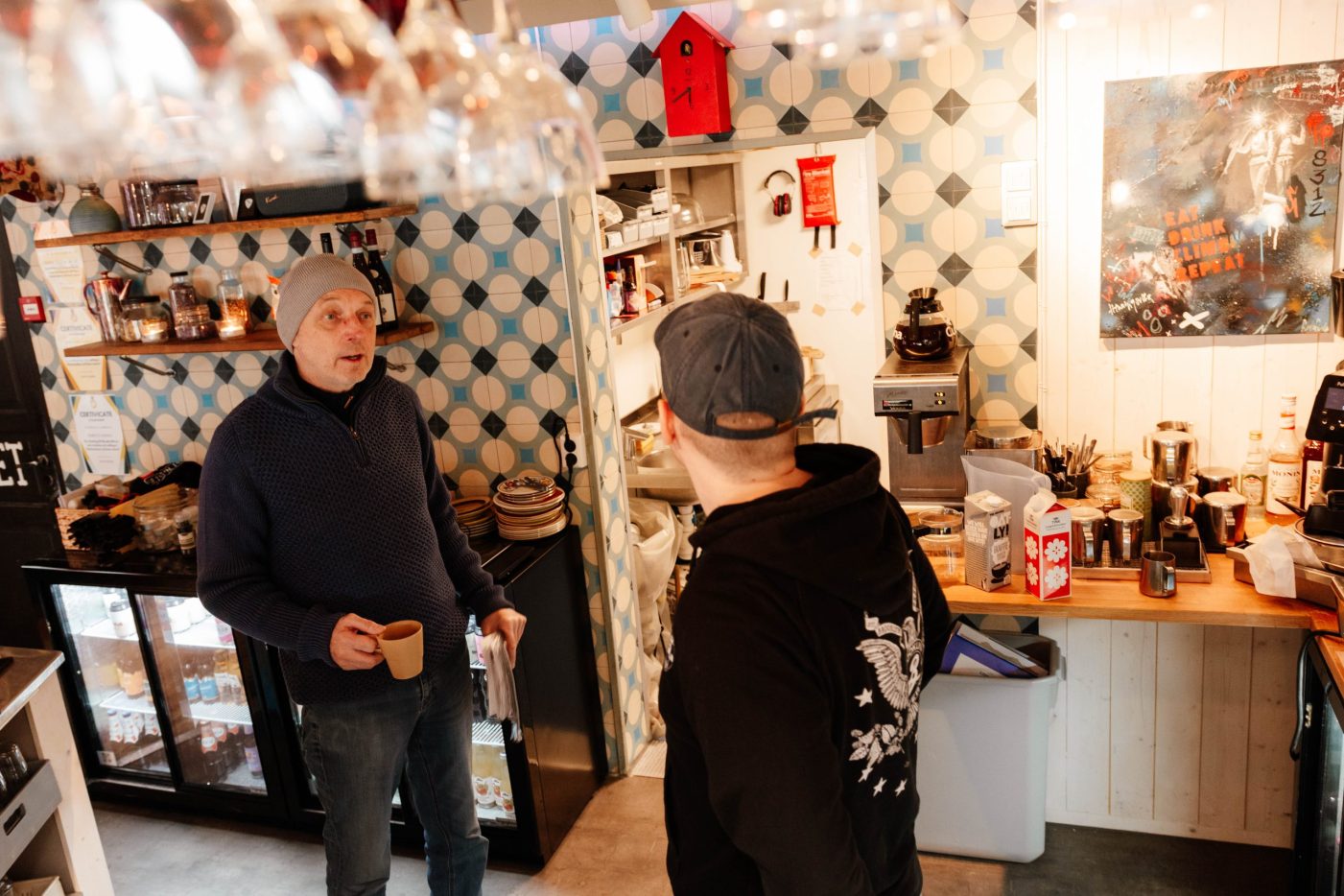 To menn i gensere og luer sitter ved et trebord på en koselig kafé, smilende og med smarttelefoner i hånden, med andre gjester og varm belysning i bakgrunnen.