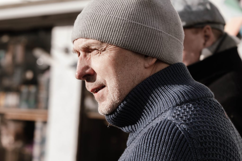 En mann iført grå strikkelue og blå genser står utendørs i sollys og ser til siden. En annen person er synlig i bakgrunnen.