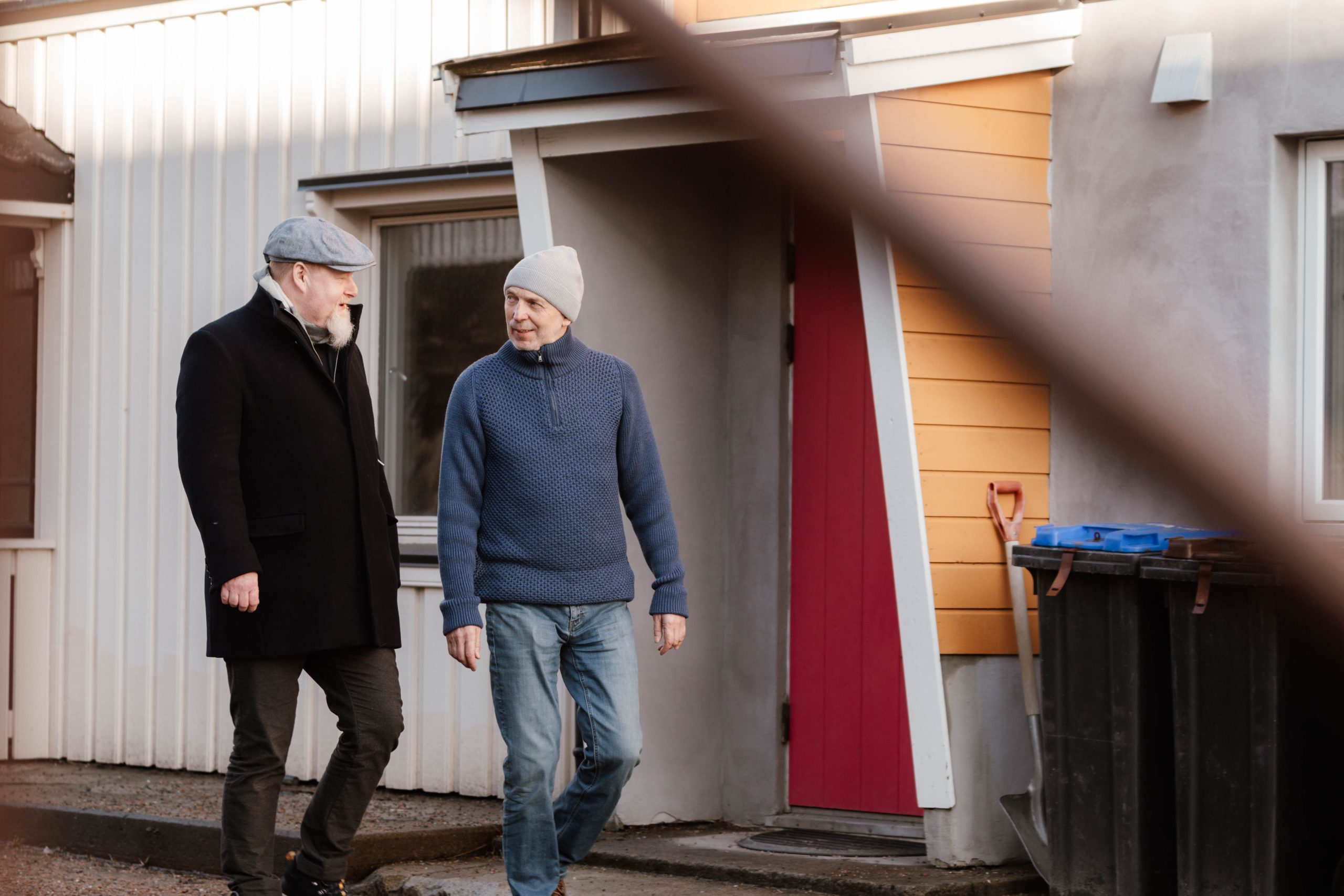 To eldre menn i frakker og hatter går og snakker utenfor et hus med en rød dør og en stor søppelbøtte i nærheten.