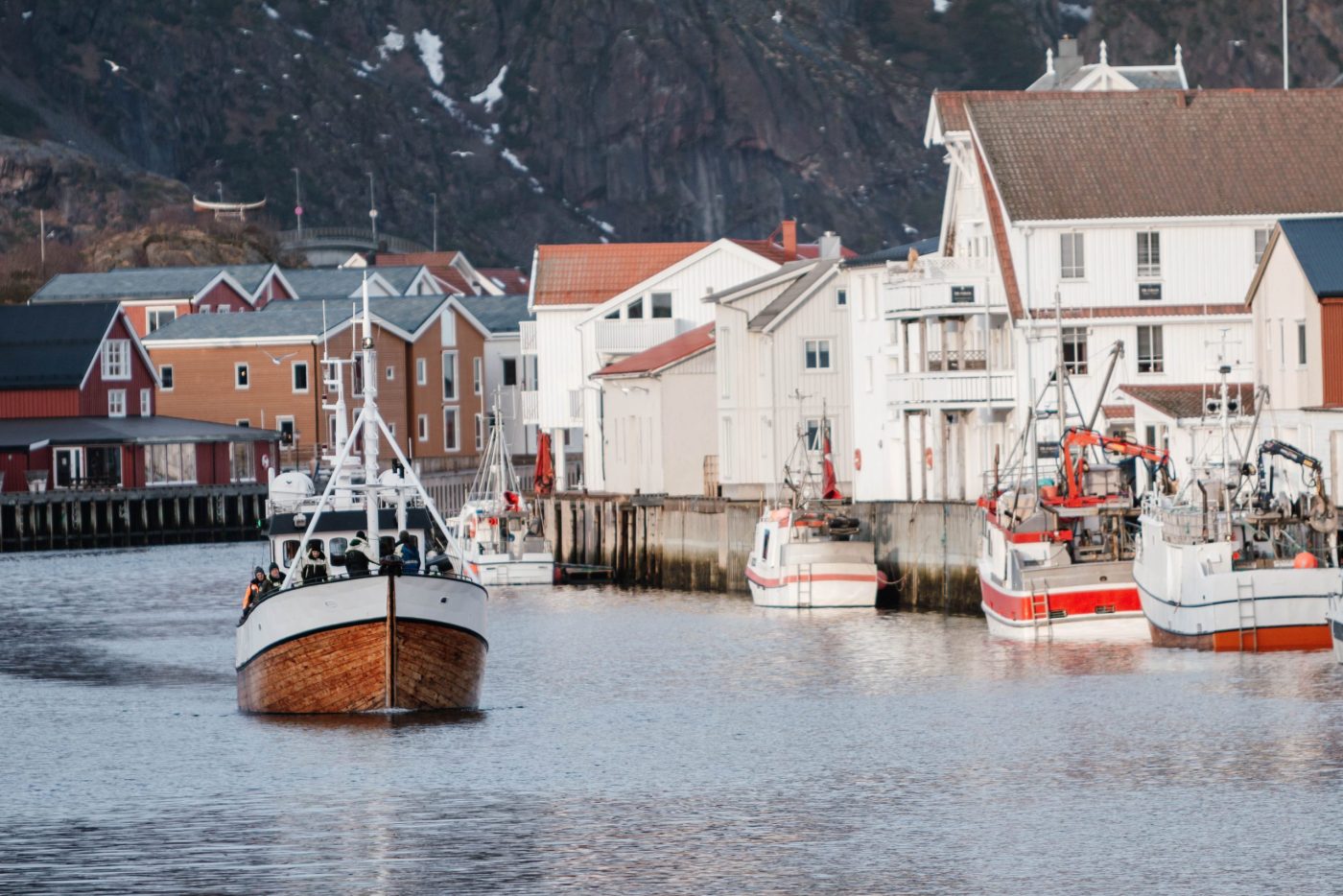 En fiskebåt av tre seiler inn mot en havn med hvite og røde bygninger og flere båter som ligger til kai, med en steinete åsside i bakgrunnen.