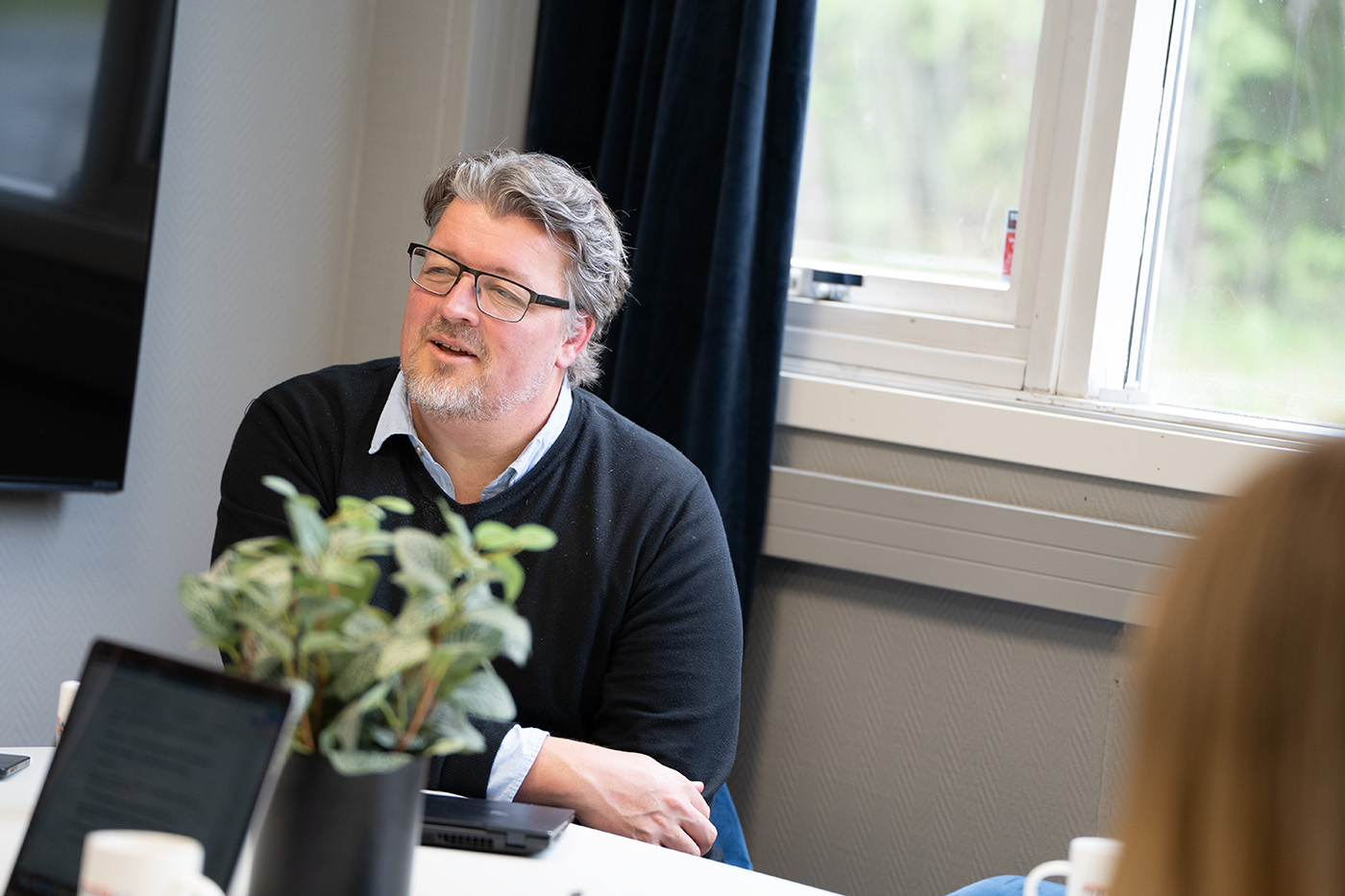 En mann med briller og grått hår sitter ved et bord på et kontor og snakker, med en bærbar datamaskin, en plante og kaffekopper foran seg.