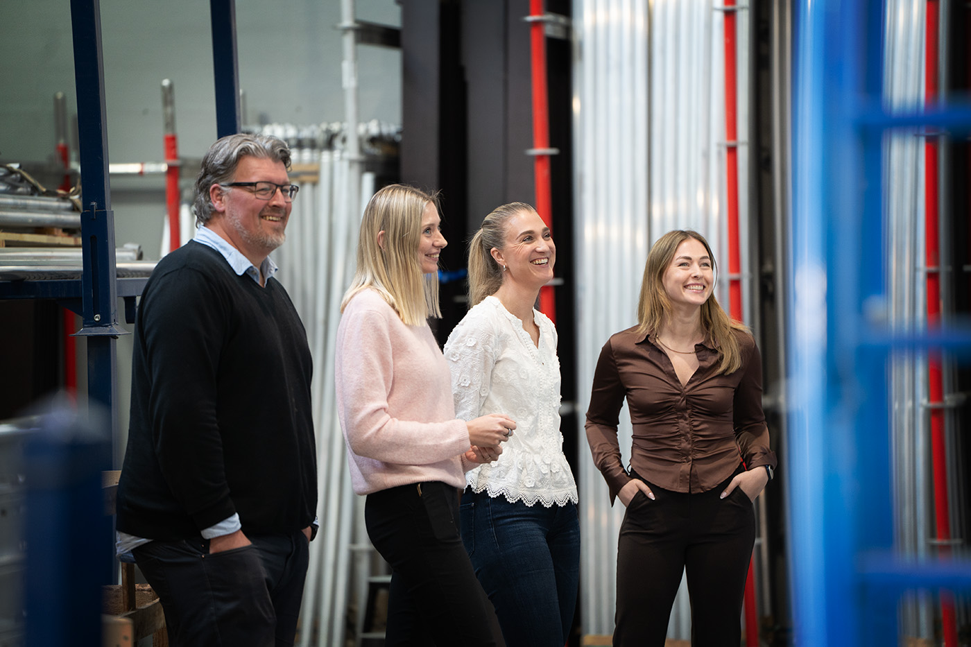 Fire voksne står innendørs, smilende og ser i samme retning, omgitt av industrirør og metallkonstruksjoner.