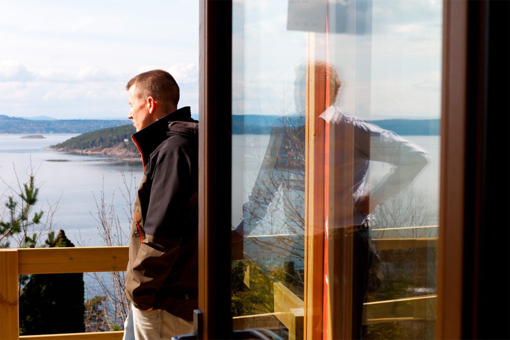 En mann står på en balkong med utsikt over vann og åser, og speilbildet hans er synlig i en glassdør.